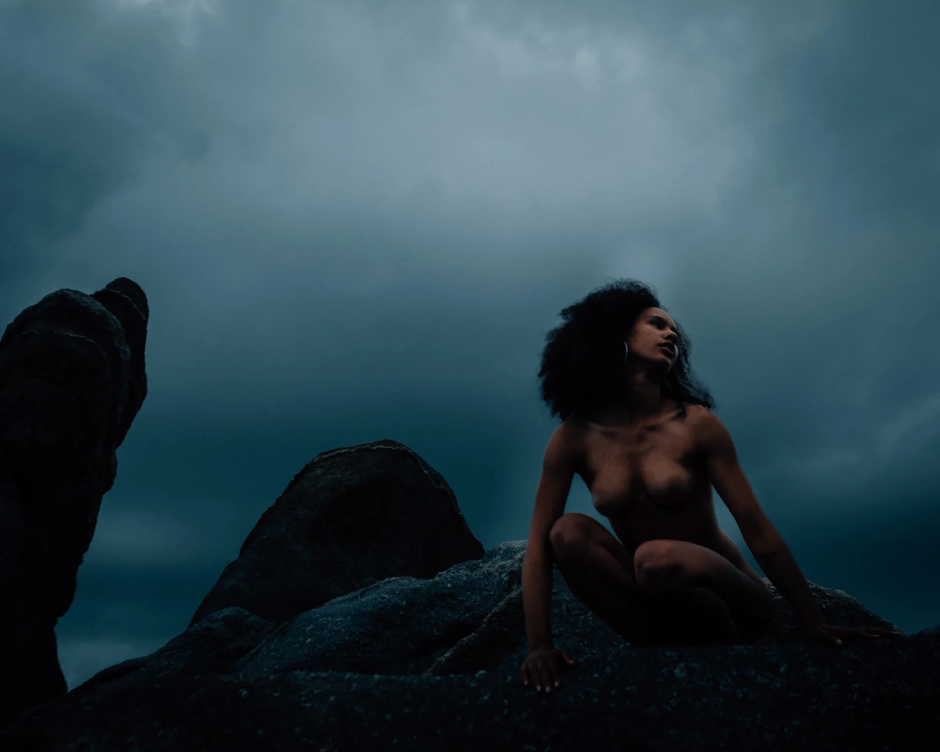 Fine art nude study — a figure seated on coastal rocks beneath a stormy sky